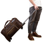 A brown leather duffle bag with wheels and adjustable long handle and multiple zipper pockets pulled by a person.