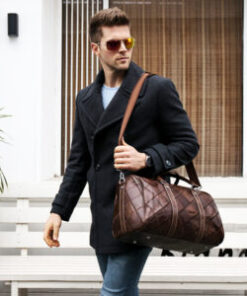A person with their face obscured carrying a brown leather travel duffle bag on his shoulder with a long shoulder strap.